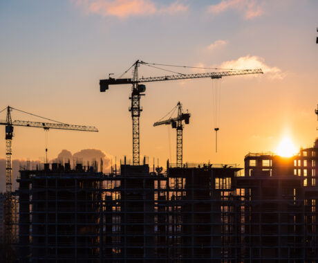 Lots of tower cranes build high-rise residential building in evening time. Steel frame structure in sunlight. Industrial background with sun flare. Yellow sunset sky.
