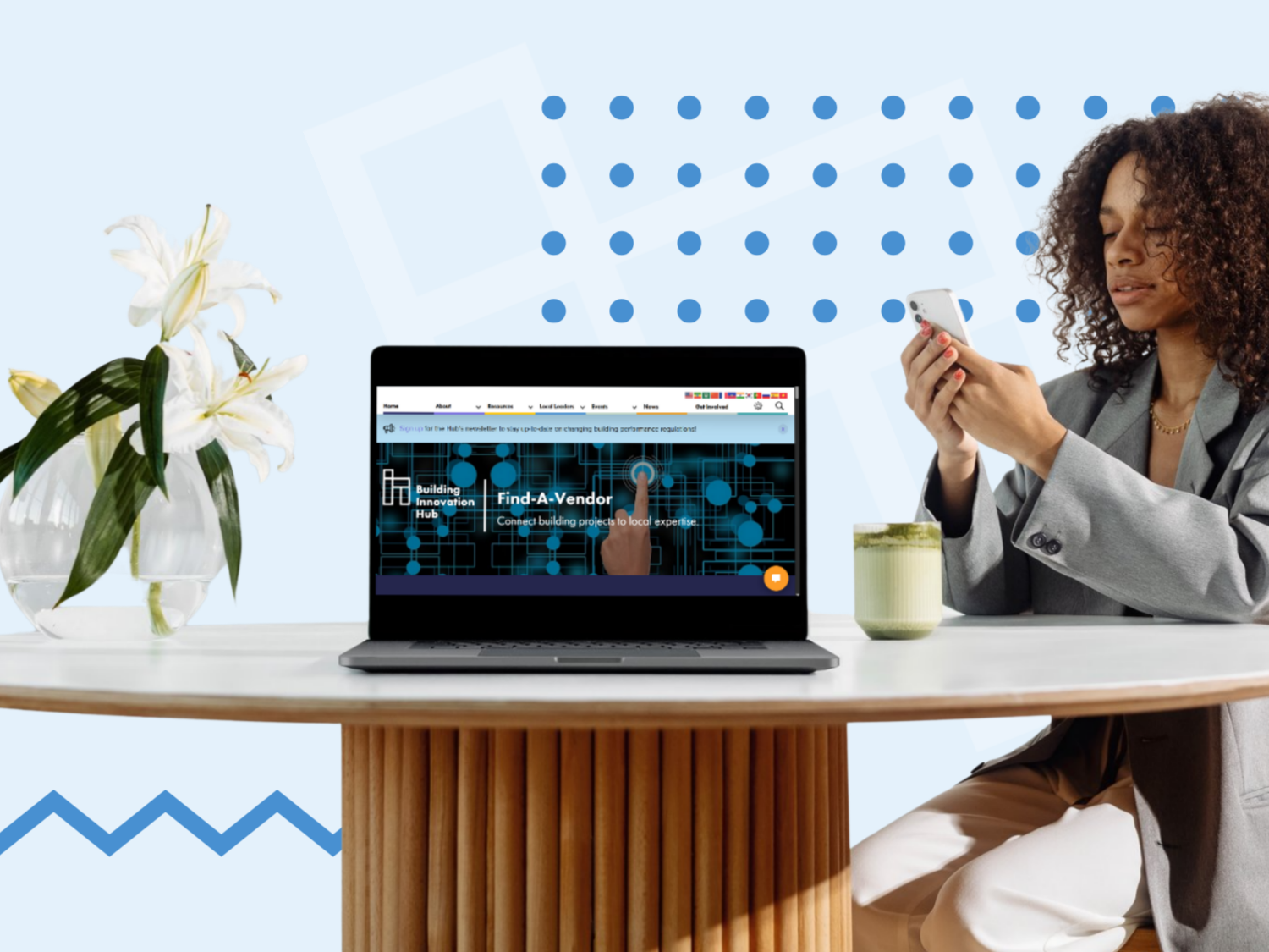 Woman in a gray suit sitting at a round table, looking at her phone, with a laptop open in front of her displaying a webpage titled ‘Find a Vendor.’ A vase with white lilies and a green mug are also on the table, with a light blue background featuring Hub-branded abstract blue shapes.